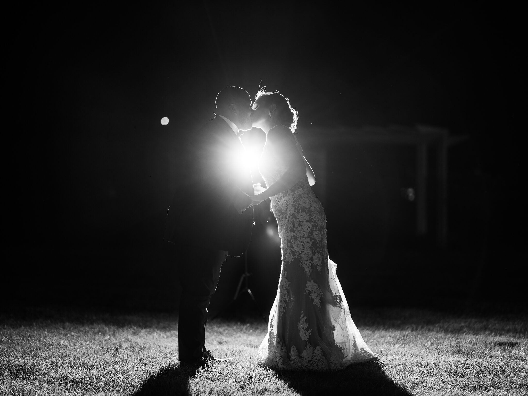 Black-and-white photo of a couple kissing, backlit by a bright light, with grass in the foreground