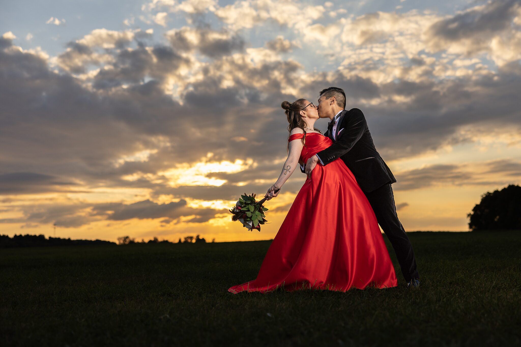 Bride in a red gown and groom share a dipped kiss in a field under a golden sunset sky