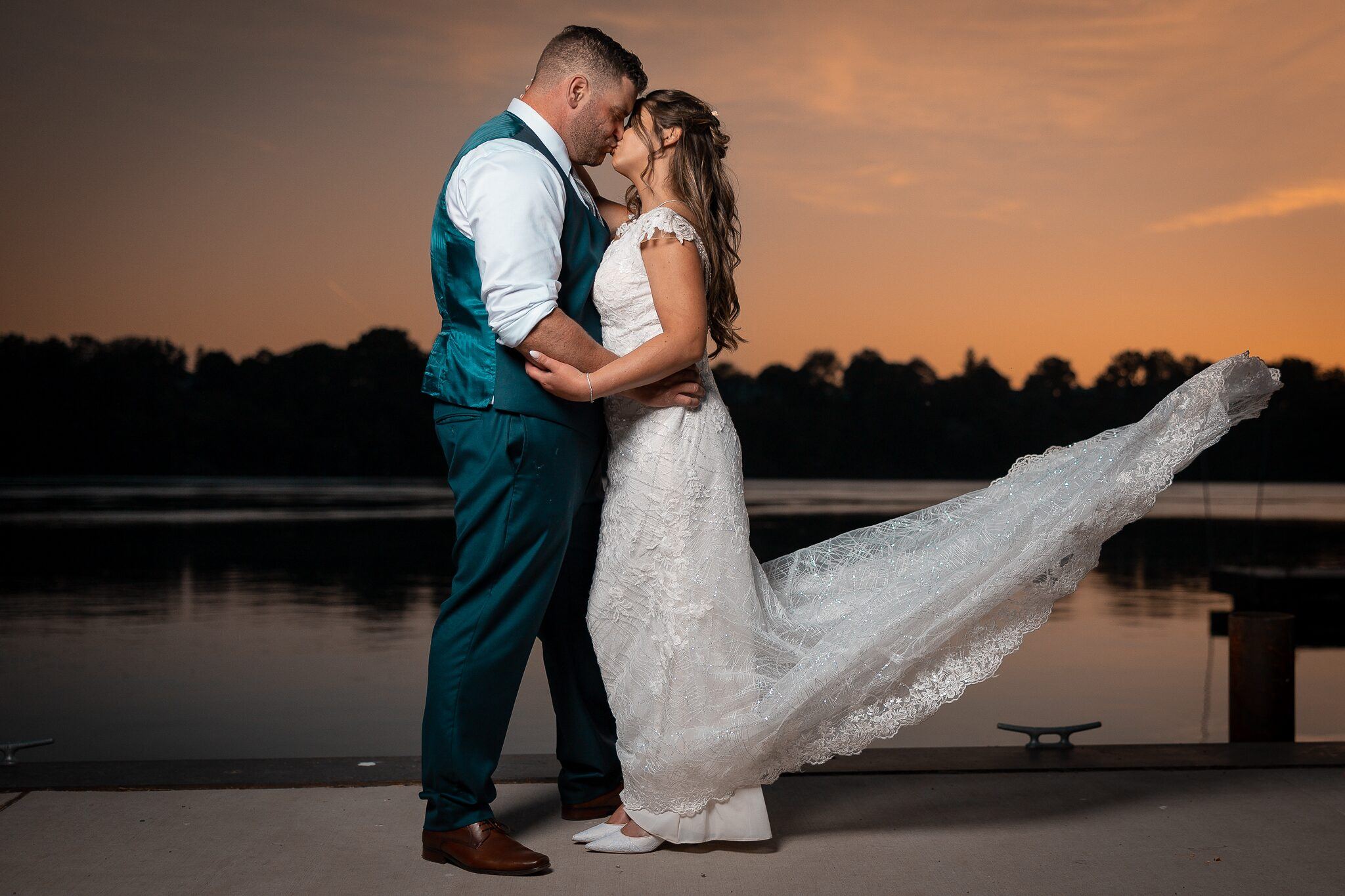 Couple embraces on a dock at twilight as the bride's lace train blows over a lake