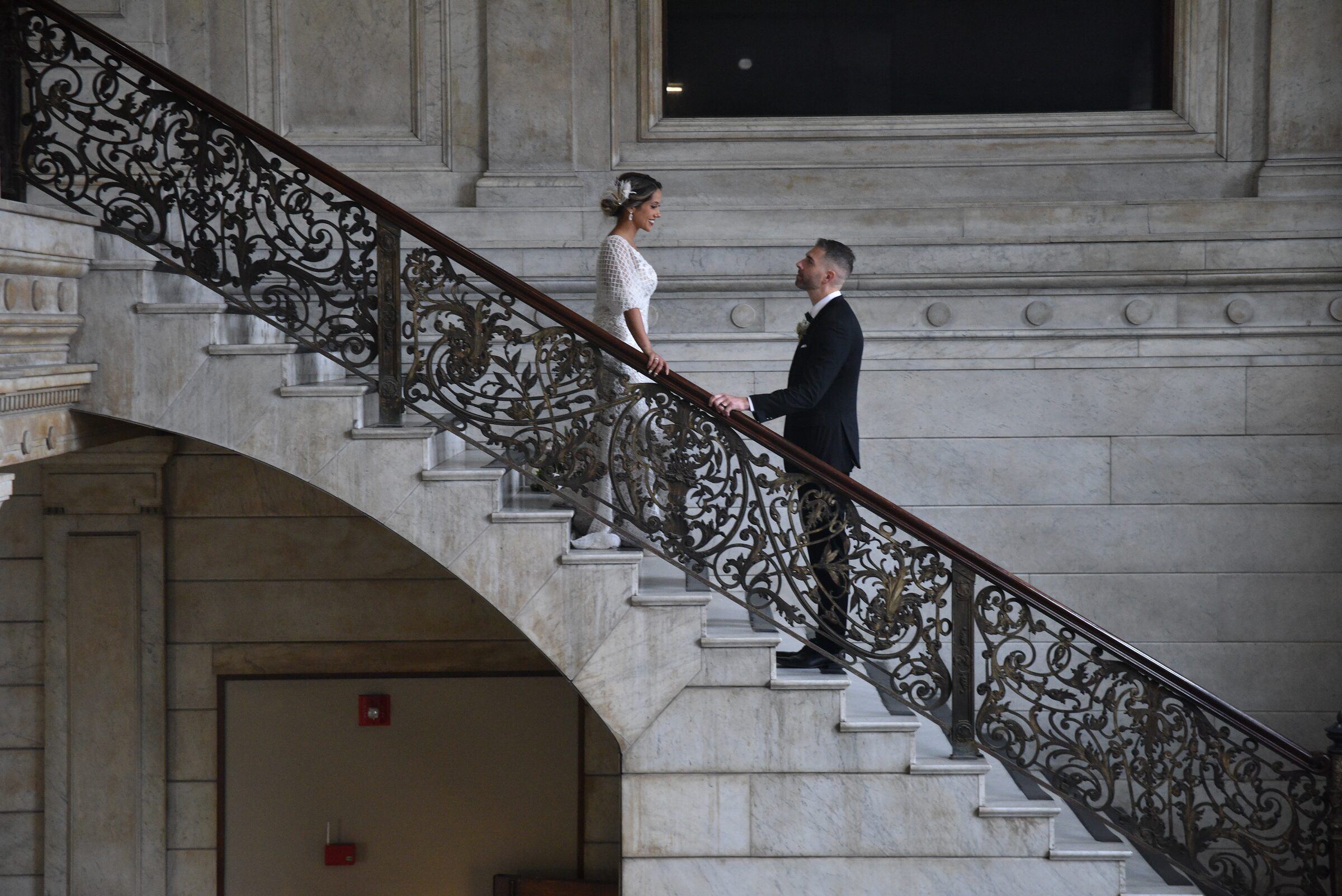 Bride and groom face each other on a grand marble staircase with an ornate iron railing