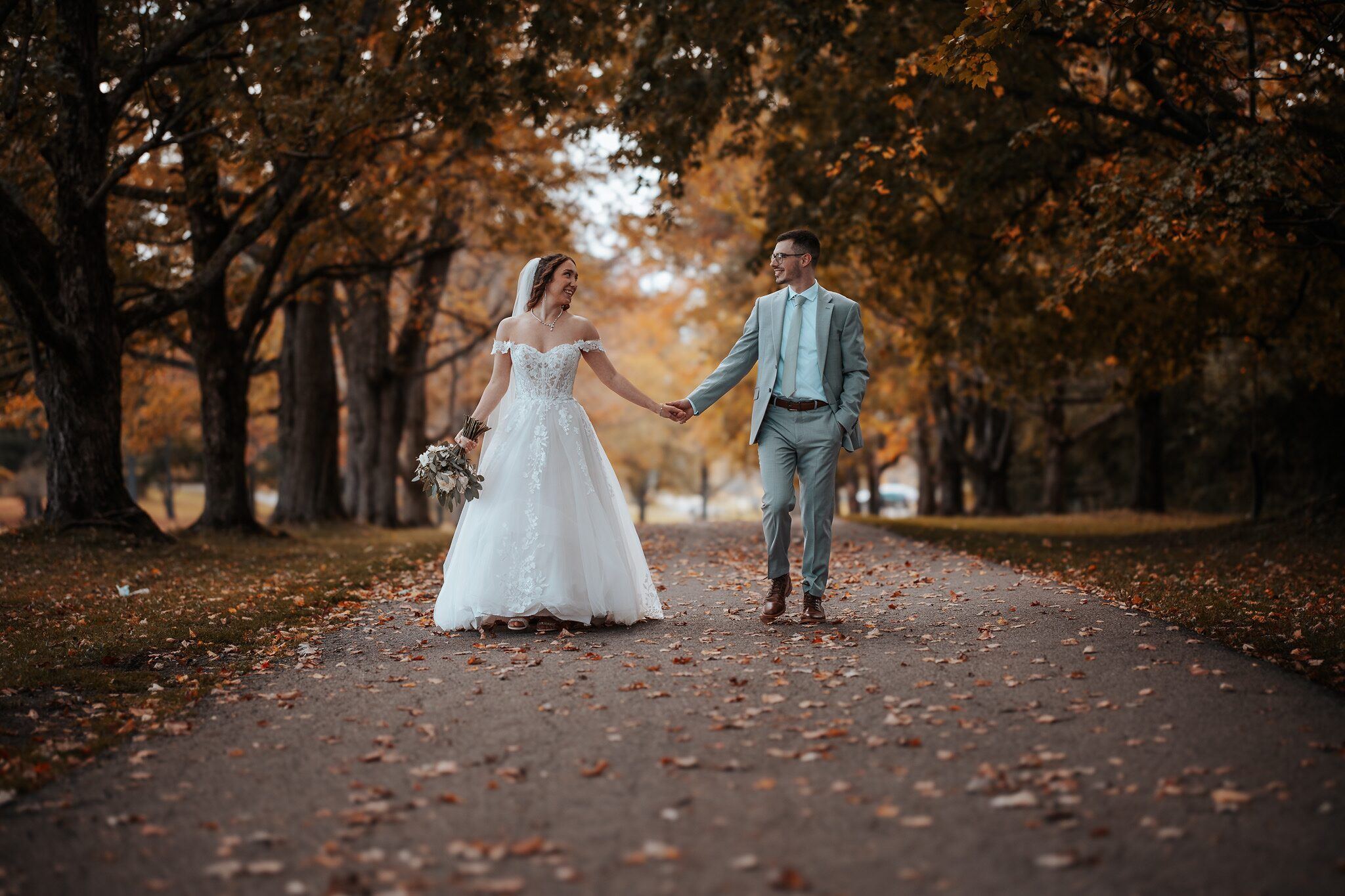Bride and groom walk hand in hand down a tree-lined path covered in fall leaves