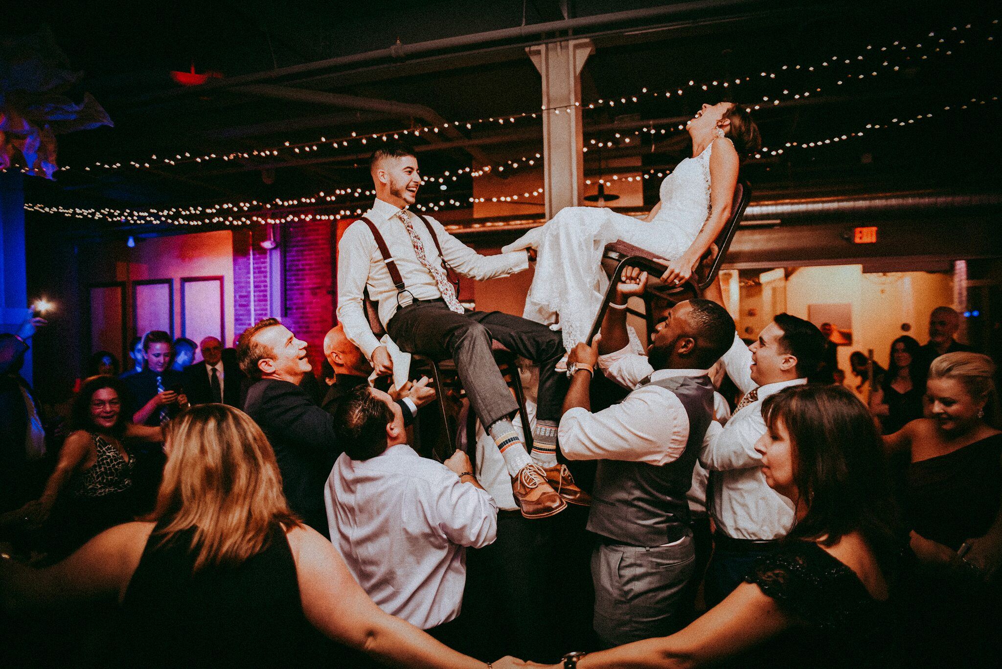Bride and groom lifted in chairs by guests during the hora at a wedding reception under string lights