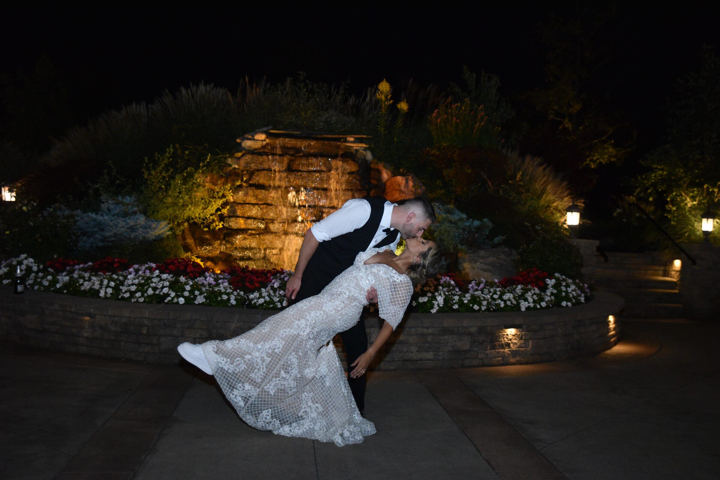 Groom dips bride for a kiss at night in a lit garden with a stone waterfall