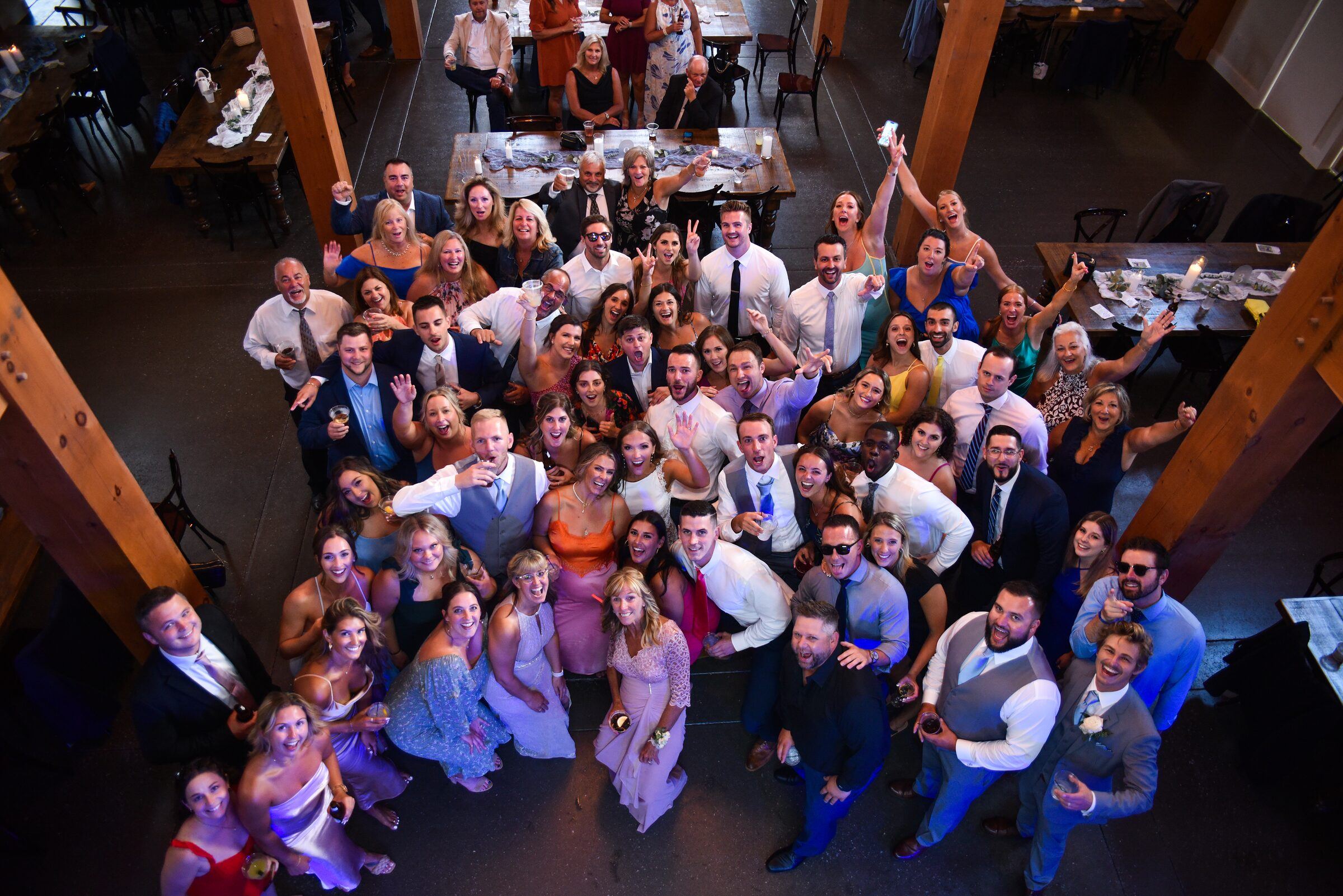 Overhead group photo of wedding guests looking up and smiling in a reception hall with wood beams
