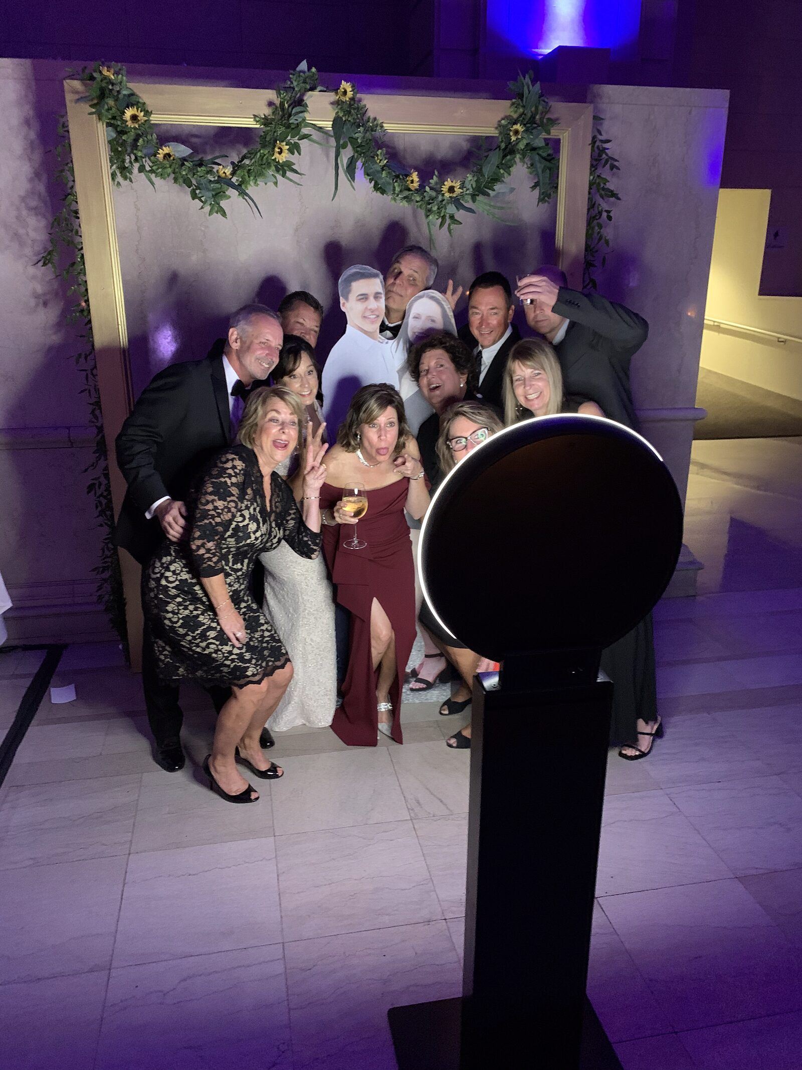 Wedding guests crowd together to pose at a round photo booth in front of a greenery-draped backdrop