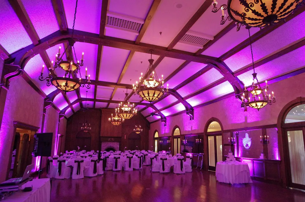 Ballroom with wood beams and chandeliers lit by magenta uplighting, tables set with white linens
