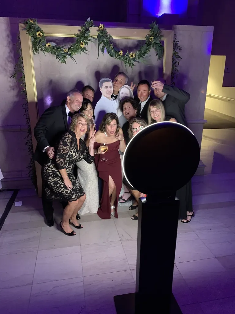 Wedding guests crowd together to pose at a round photo booth in front of a greenery-draped backdrop